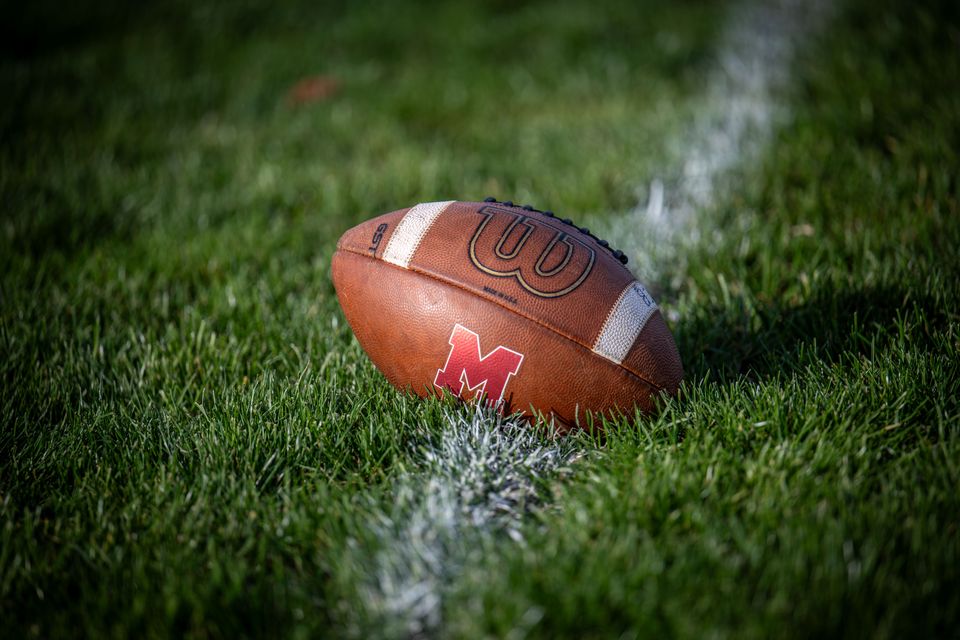 A football with a red M on it resting in the grass beside a white yard line.