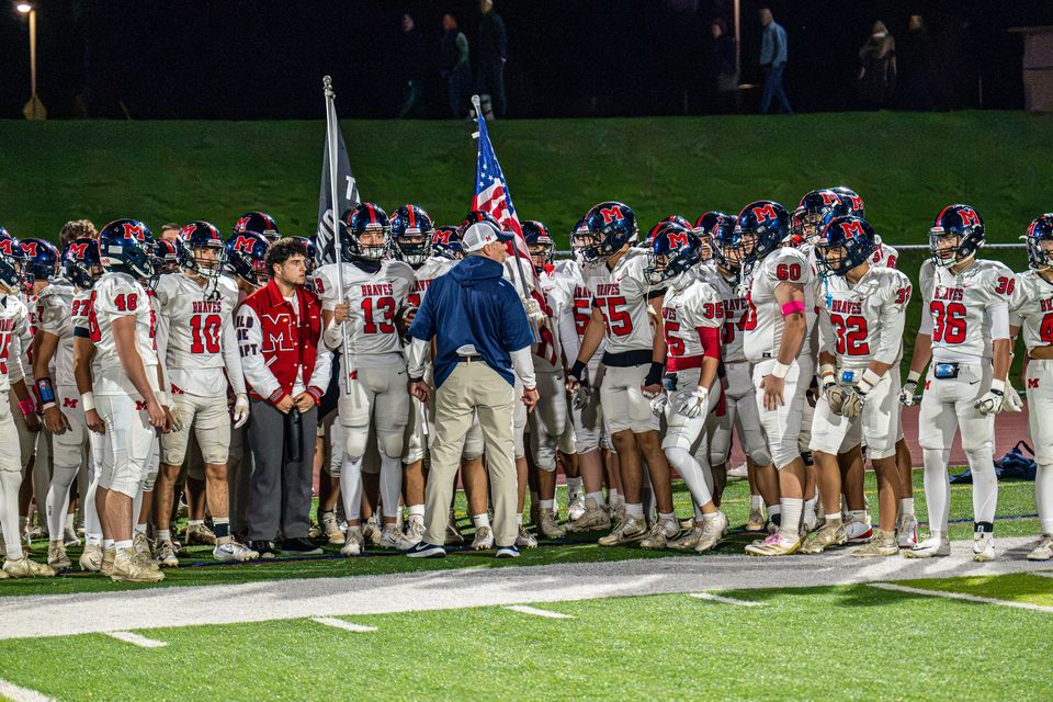 The Manalapan Braves gather on the sideline under the lights, with the team flags behind them.