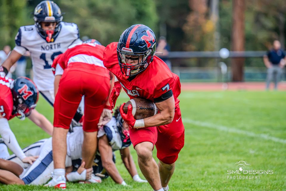 A Manalapan running back in red drives forward with the ball past a pile of players.